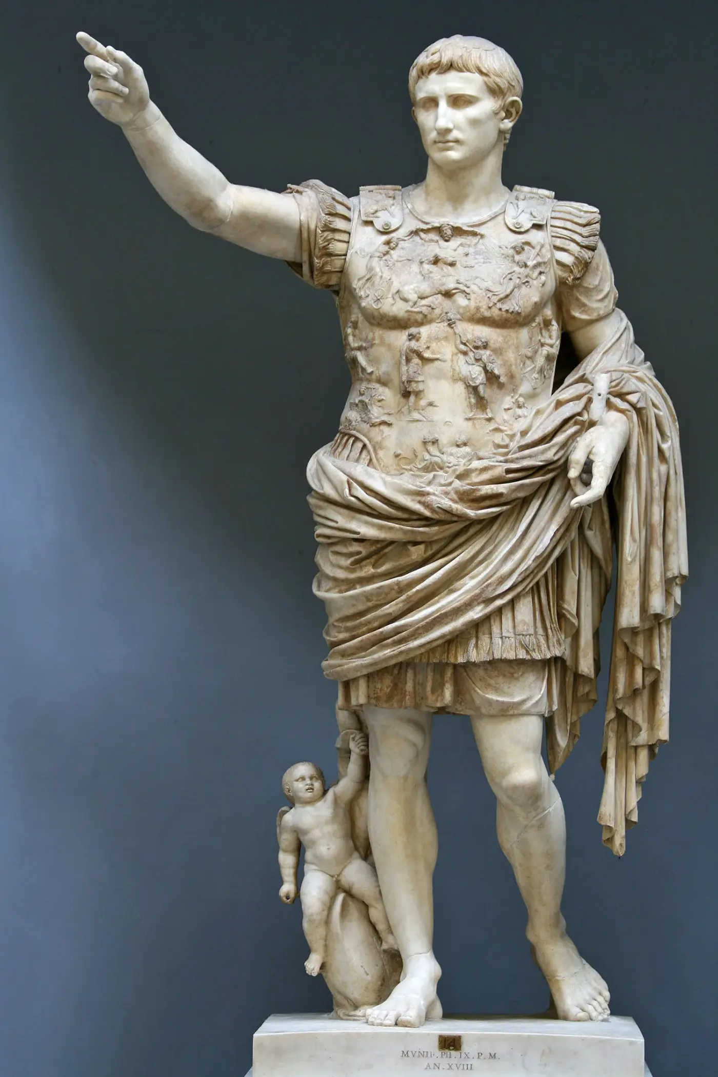 Estatua de mármol de un emperador romano en pie, con coraza decorada en relieve, el brazo derecho alzado en gesto de arenga y un pequeño cupido junto a la pierna derecha.